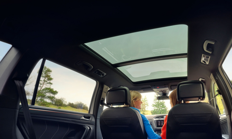 Side view of the front of a red Tiguan Allspace, in the background a woman with three children is standing at the fence of a horse pasture. 