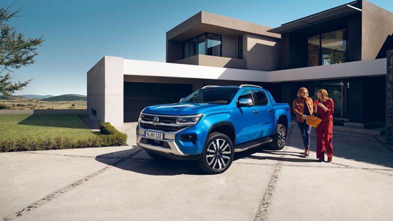  Un pick-up Volkswagen Amarok bleu est garé devant une maison moderne. Deux femmes marchent sur l'allée à côté du véhicule.