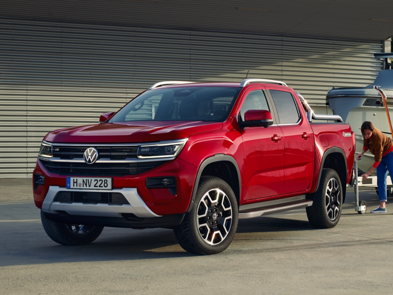 Vor einer grauen Gitterfassade steht der rote Volkswagen Amarok mit silbernem Stoßfänger, während eine Frau im Hintergrund den angekoppelten Bootstrailer positioniert.