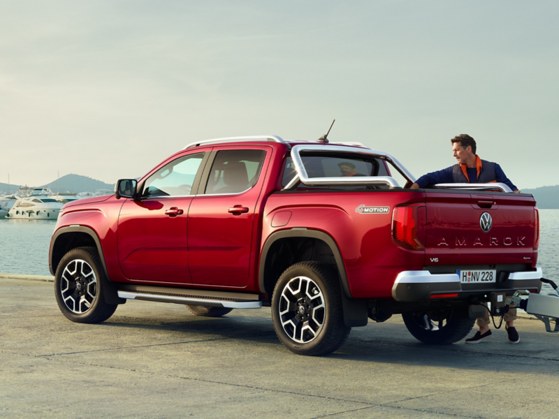 Roter VW Amarok steht an einem Kai mit Yachten im Hintergrund, während ein Mann im Anzug über die geöffnete Ladeklappe des Trucks schaut, der einen Anhänger zieht.