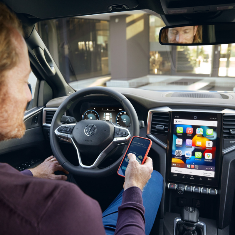 Un homme est assis sur le siège conducteur d'une VW Amarok et tient un smartphone à la main. L'écran du véhicule affiche les icônes de diverses applications de connectivité.