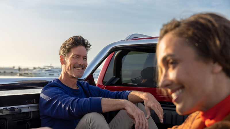 Ein Mann und eine Frau sitzen auf der Ladefläche eines VW Amarok, im Hintergrund sieht man Schiffe