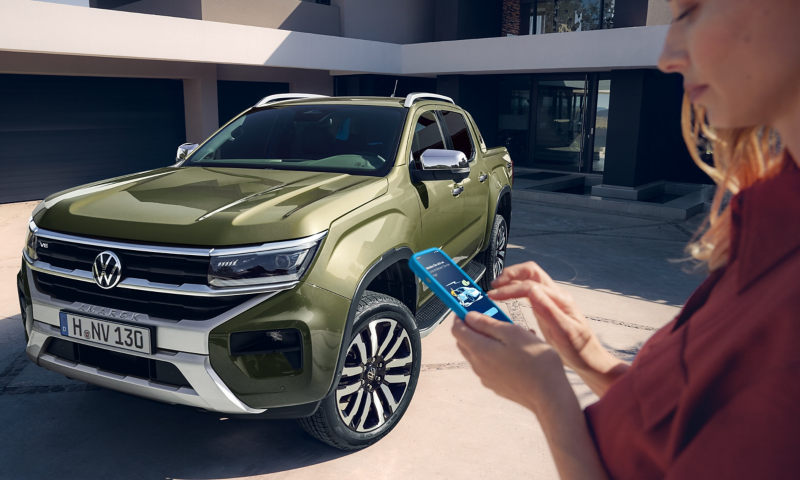 Ein VW Amarok steht in einer Einfahrt vor einem modernen Haus und eine Frau im Vordergrund schaut in ihr Smartphone.