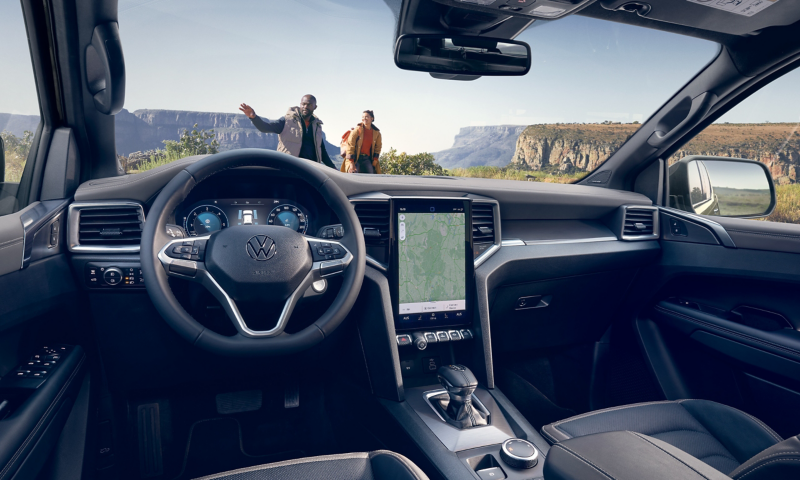 Der Innenraum eines VW Amarok, welcher vor einer bergigen Landschaft steht. Vor dem Fahrzeug stehen zwei Personen.