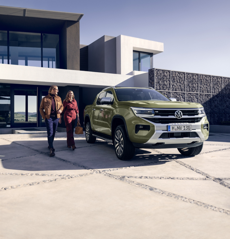 Una pick-up Volkswagen Amarok de color verde oliva estacionada frente a una moderna casa con un diseño arquitectónico elegante. Una pareja camina hacia el vehículo por un camino empedrado, sugiriendo un estilo de vida contemporáneo y práctico.