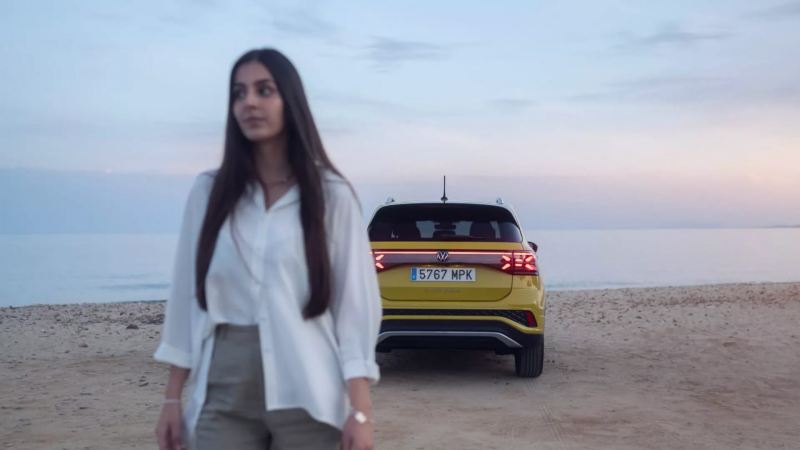 Una chica en primer pano y un Volkswagen T-Cross visto desde atrás en la playa, Volkswagen Approved