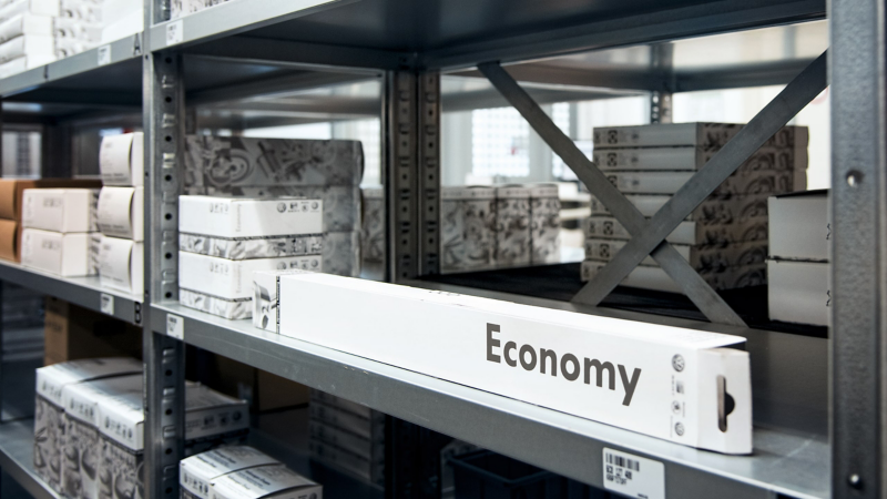 Une longue boîte blanche étiquetée "Economy" est posée sur une étagère métallique dans un entrepôt, avec d'autres boîtes et colis empilés sur les étagères en arrière-plan.