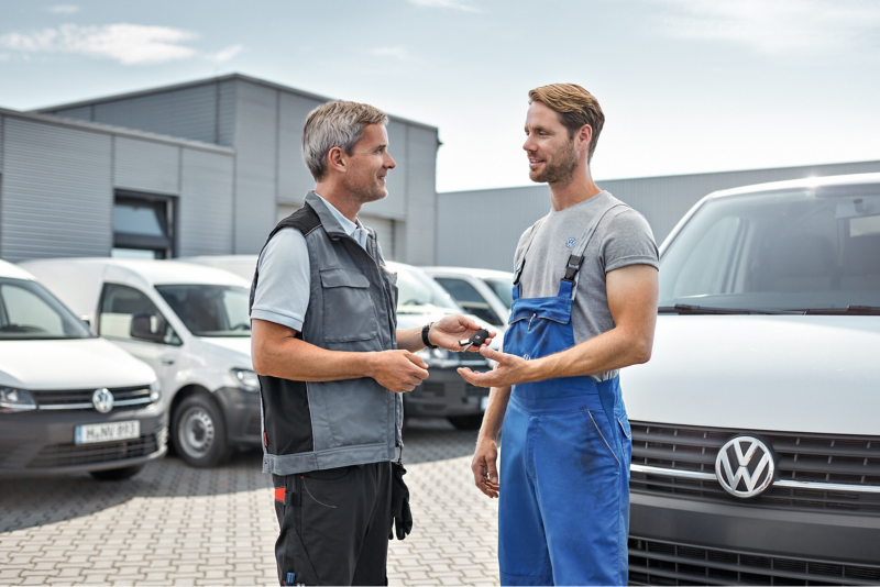 Un asesor de servicio con un chaleco gris entrega las llaves de un vehículo a un técnico vestido con mono de trabajo azul, frente a una fila de furgonetas Volkswagen blancas en un centro de servicio. La imagen simboliza la entrega o recogida de un vehículo en el taller.