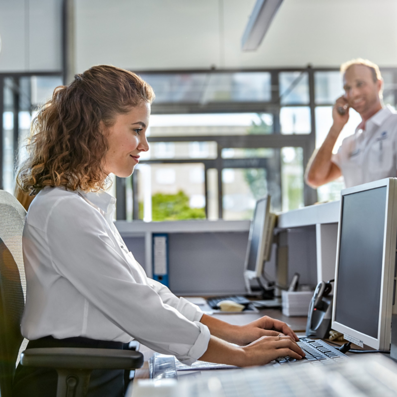 Eine Frau in weißer Bluse sitzt in einem Büro und arbeitet, im Hintergrund telefoniert ein Mann.