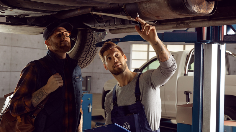  Deux hommes se tiennent sous un véhicule soulevé dans un garage. L'un, portant des lunettes et une veste, regarde le véhicule, tandis que l'autre, en salopette, pointe du doigt un élément sous la voiture.