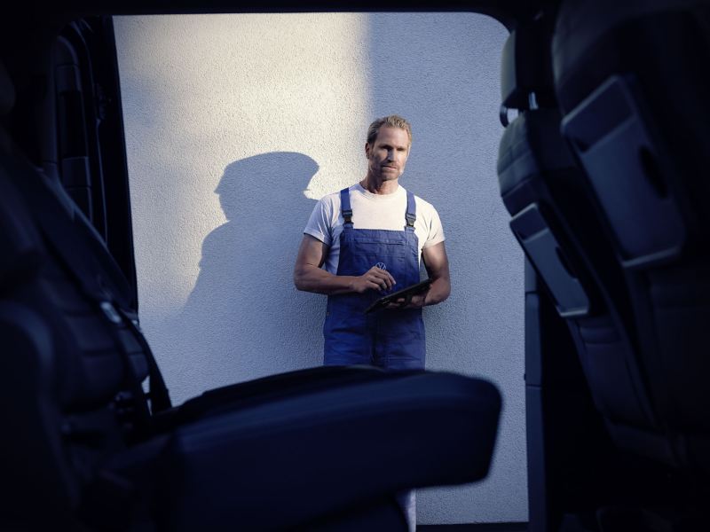 Ein Mann mit Clipboard steht neben einem hochgebockten VW Nutzfahrzeug in einer Werkstatt.