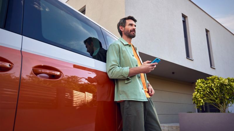Ein Mann mit Smartphone lehnt an einem VW Multivan.