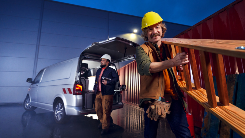 Ein Handwerker lädt eine Leiter aus einem VW Fahrzeug aus, vor einer Lagerhalle.