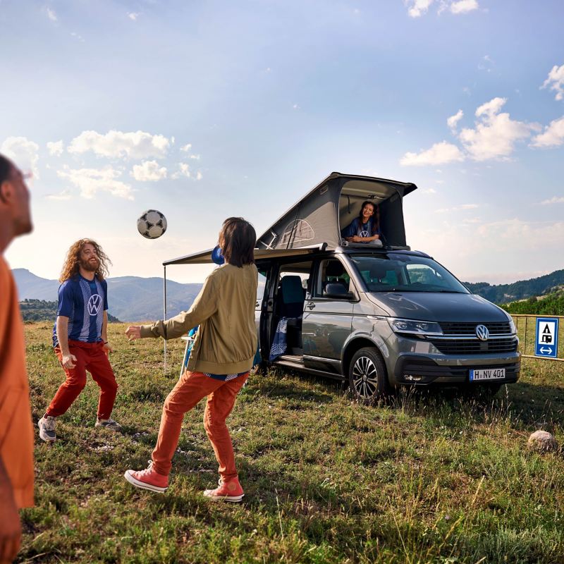 Freunde spielen vor einem VW California 6.1 mit aufgestelltem Aufstelldach Fußball.