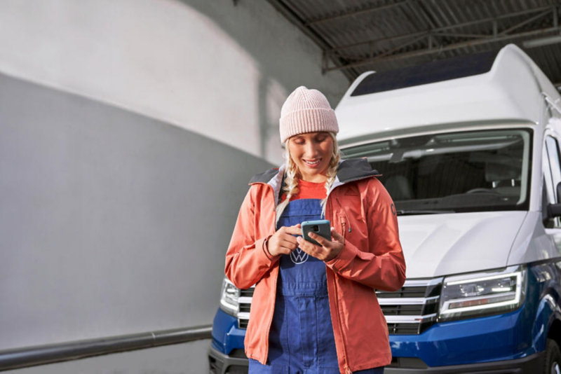 Une technicienne sourit en consultant son smartphone, debout dans un garage devant un vw Grand Callifornia Volkswagen.