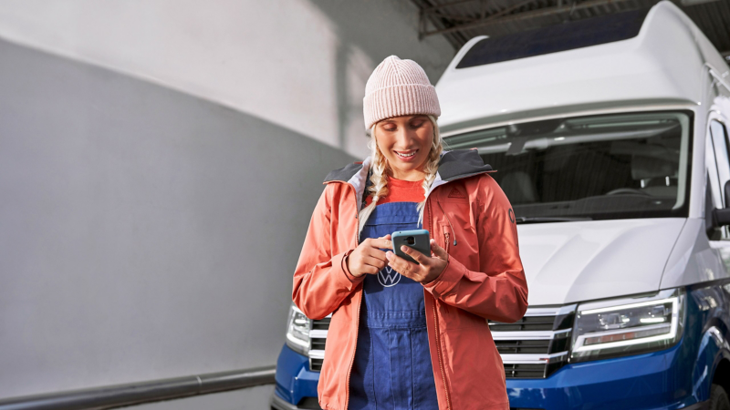 Junge Frau in Arbeitskleidung und Mütze lächelt und benutzt ihr Smartphone vor einem Volkswagen Campervan. 