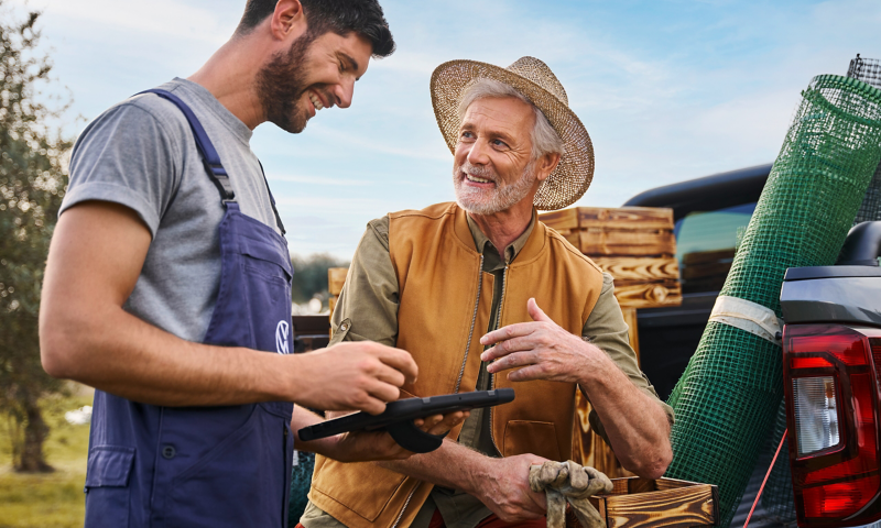 Ein VW Servicemitarbeiter steht bei einem älteren Herren an seinem VW Nutzfahrzeug in der Natur und hält ein Tablet in der Hand.