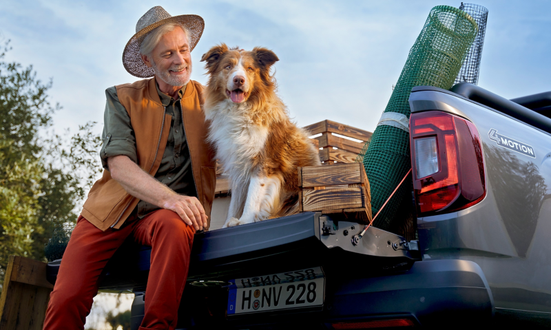 Ein älterer Herr sitzt mit seinem Hund auf der Ladefläche seines VW Amarok in der Natur.