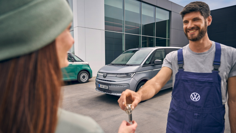 Ein lächelnder Volkswagen-Mitarbeiter in blauer Arbeitslatzhose übergibt einer Kundin vor einem modernen Autohaus die Fahrzeugschlüssel für einen silbernen VW Multivan.