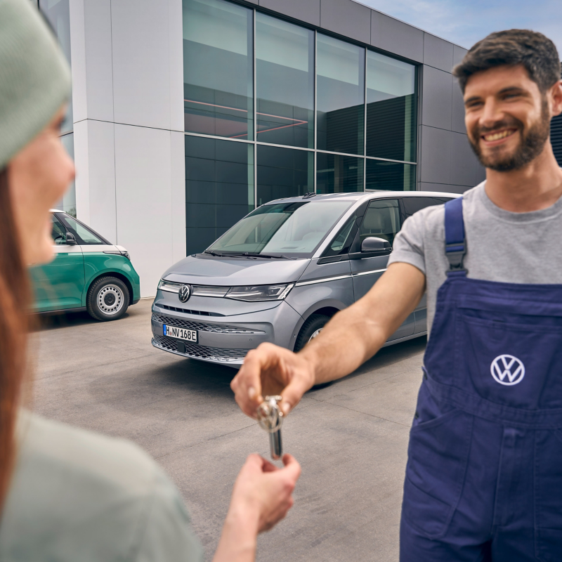 Ein lächelnder Volkswagen-Mitarbeiter in blauer Arbeitslatzhose übergibt einer Kundin vor einem modernen Autohaus die Fahrzeugschlüssel für einen silbernen VW Multivan.