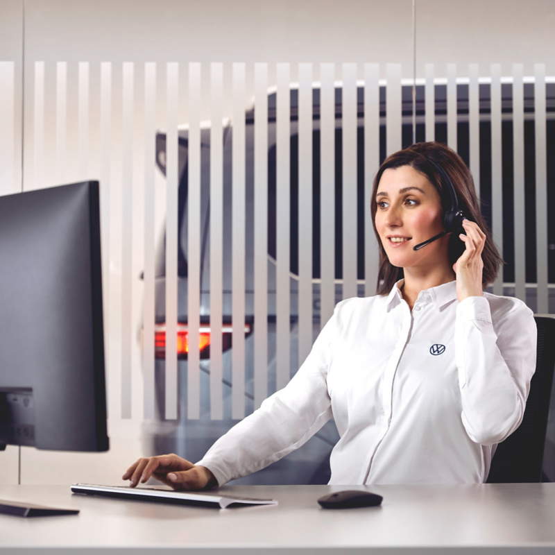 Eine Frau in weißer Bluse sitzt in einem Büro und arbeitet, im Hintergrund sieht man ein VW Nutzfahrzeug stehen.