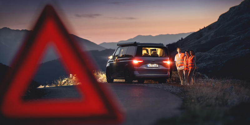 Ein Warndreieck im Vordergrund, eine Reisegruppe mit liegengebliebenem VW Nutzfahrzeug bei Dämmerung wartet im Hintergrund auf den Pannendienst