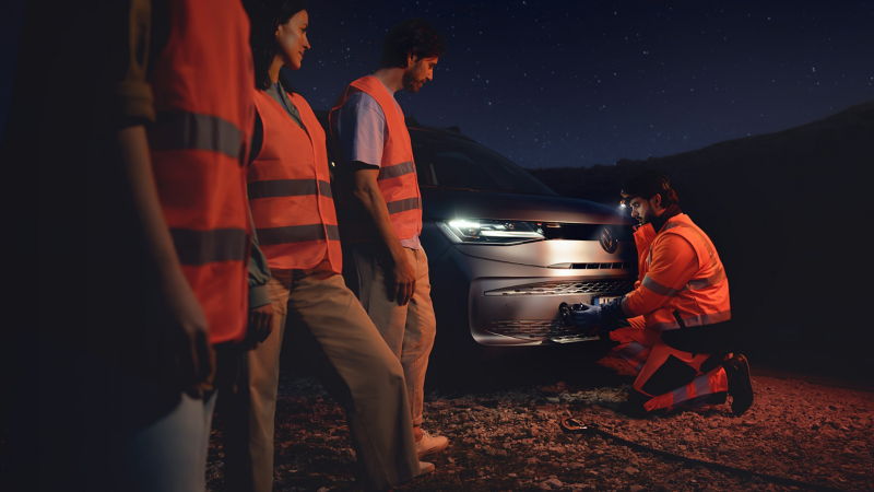 Ein Pannenhelfer repariert ein VW Nutzfahrzeug bei Nacht, die Insassen warten daneben in Warnwesten