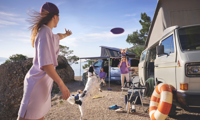 Une famille s'amuse sur un campement en bord de mer, entre un Volkswagen Combi T3 classique et un van California moderne. Une femme lance un frisbee qu'un chien essaie d'attraper en sautant.