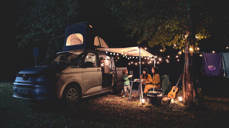 En VW California i skymningen – en familj sitter vid ett bord under markisen, omgivna av varma ljusslingor.