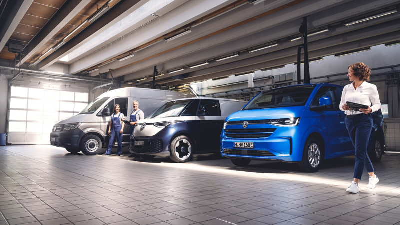 Drei Volkswagen-Transporter stehen in einer Werkstatthalle, während eine Frau mit einem Tablet und ein Paar Mechaniker daneben stehen.