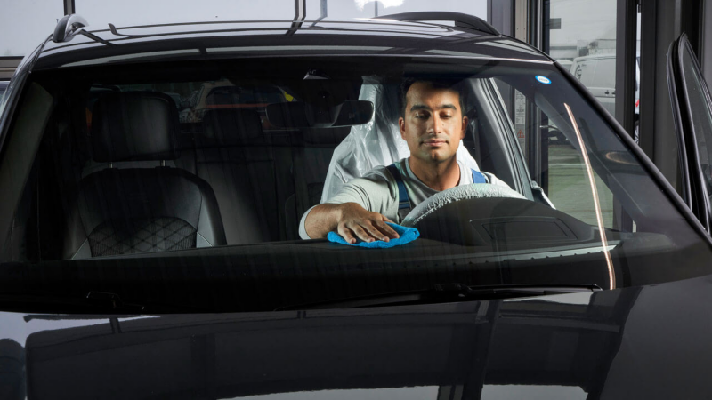 Un technicien Volkswagen nettoie l'habitacle d'une voiture, vue à travers le pare-brise.