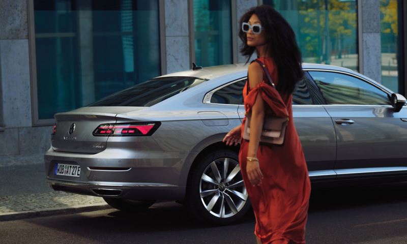 Seitlicher Blick auf das Heck eines silbernen VW Arteon mit eHybrid-Badge, im Vordergrund eine Frau im Kleid mit Sonnenbrille.