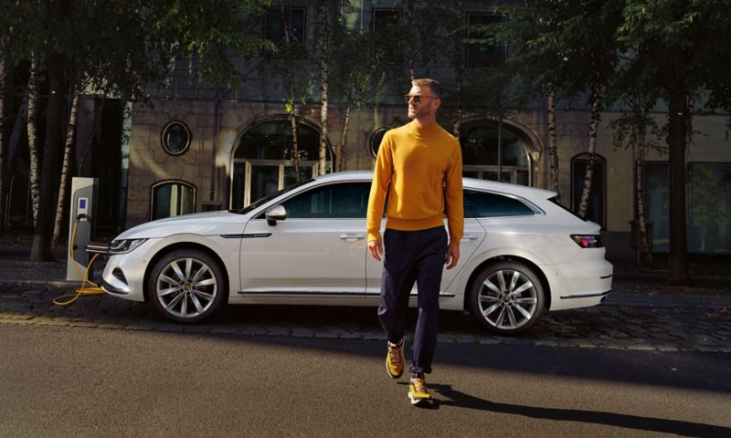 En VW Arteon Shooting Brake eHybrid holder i vejsiden og lades op ved en ladestander i urbane omgivelser, en mand iført solbriller går væk fra bilen.