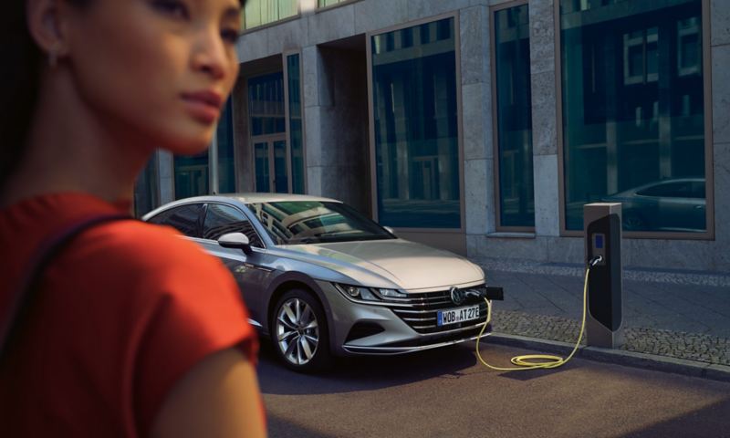 En sølvfarvet VW Arteon eHybrid holder foran en moderne kontorbygning og bliver ladet op ved en ladestander, i forgrunden går en kvinde forbi.