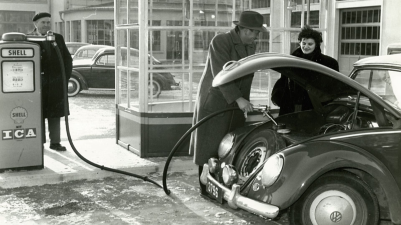 Photographie en noir et blanc d'une Volkswagen Coccinelle en train de faire le plein à une pompe Shell vintage, avec des personnes à proximité.