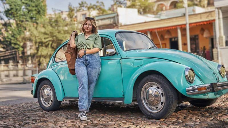 Joven mujer posa delante de un Vocho de Volkswagen en color azul turquesa. 