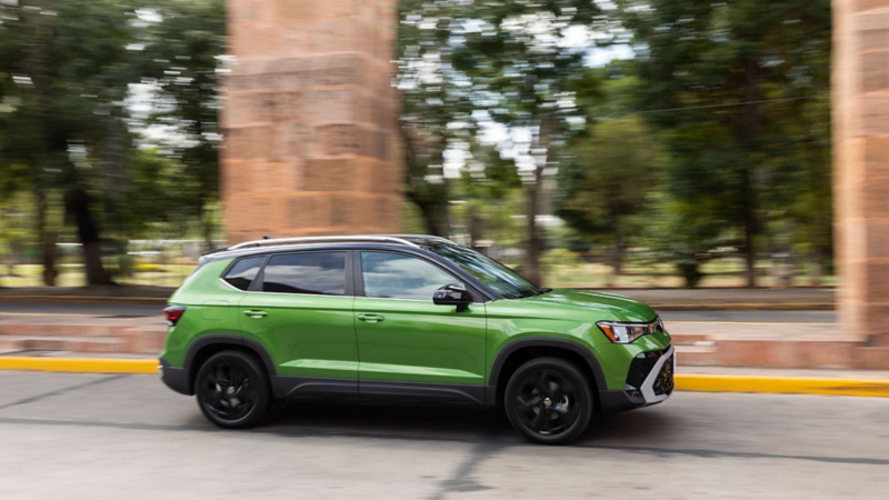 SUV verde que recorre una avenida arbolada junto a una acera urbana.