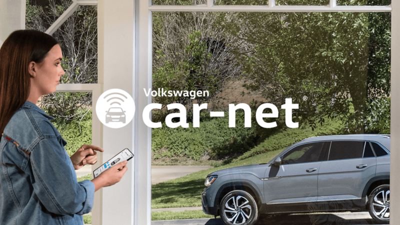 A woman looking at a 2022 Volkswagen Atlas Cross Sport SUV through a window while holding a mobile phone with the Car-Net® app on the screen.