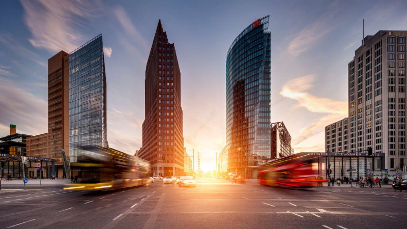 Paysage urbain dynamique au coucher du soleil, avec de hauts bâtiments modernes et des véhicules (bus rouges et jaunes) filant sur une route, créant un effet de flou de mouvement. Le soleil éclaire le centre de l'image, possiblement à Potsdamer Platz.