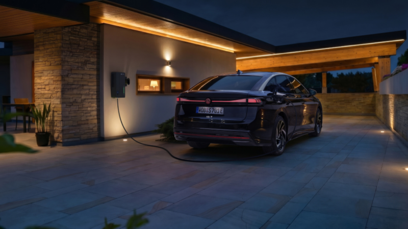 A dark-colored electric sedan is parked at night on a modern paved driveway, plugged into a wall charger mounted on a house with warm exterior lighting.