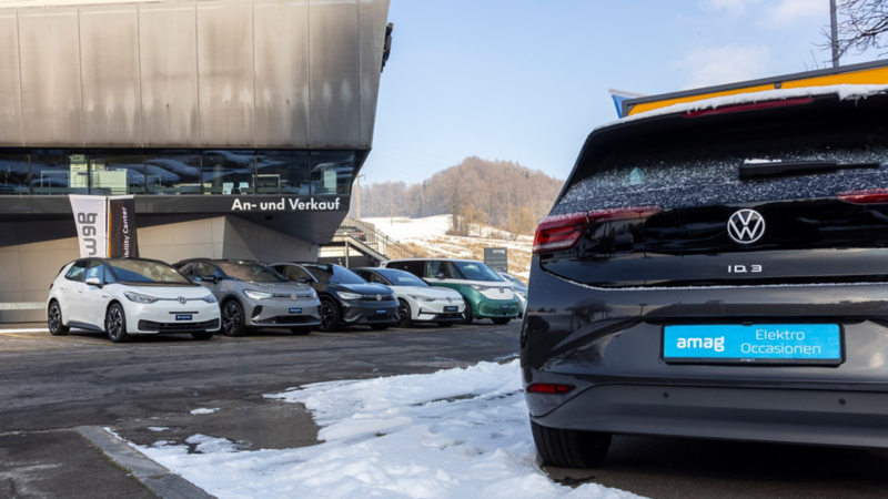 Posteriore di una Volkswagen ID.3 in primo piano, con diverse auto elettriche VW usate davanti all’edificio e neve a terra.