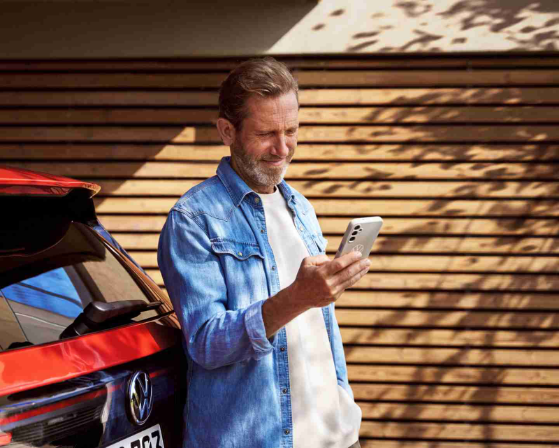 Un hombre de mediana edad con barba y camisa vaquera, de pie junto a un auto rojo mientras mira su teléfono inteligente.