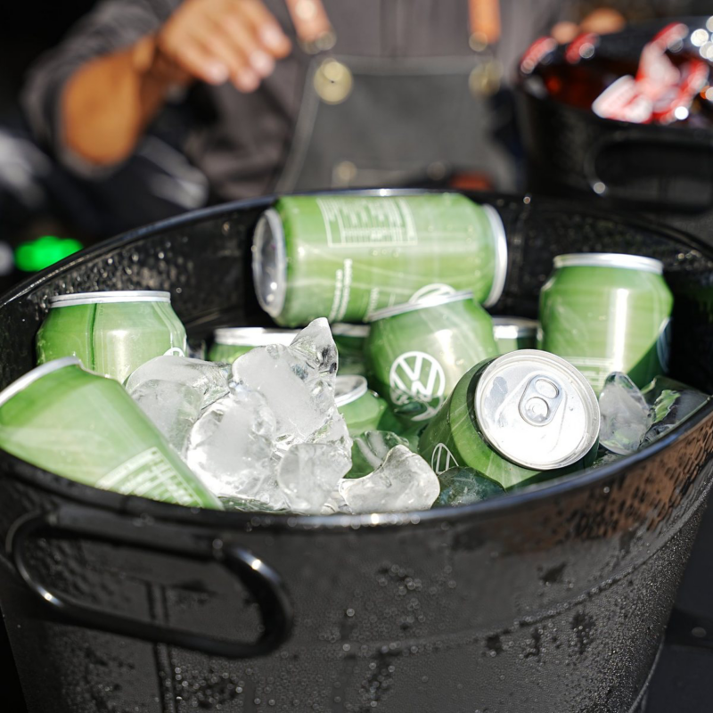 Cubeta negra con hielo y latas verdes de bebida, con una persona parcialmente visible al fondo.