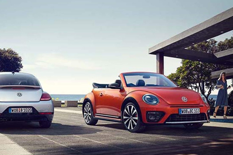 A orange Volkswagen Beetle Cabriolet, parked next to a beach on a promenade.