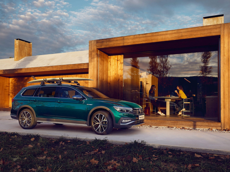 A green Volkswagen Passat Estate, with a surfboard on the roof-rack. Parked outside a glass-fronted beach house.