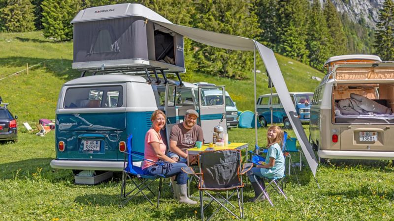 blauer Bulli parkiert auf der Wiese und eine Familie sitzt mit dem Campingtisch davor