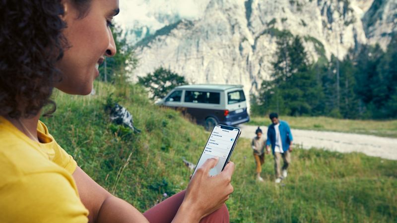 Eine Frau bedient im Vordergrund die Standheizung auf einem Smartphone, im Hintergrund sind ein Mann, ein Kind und ein VW California zu sehen.