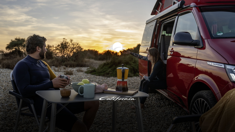Ett par äter frukost i campingmöblerna utanför en röd VW California med taket uppfällt.