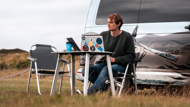 En man sitter och jobbar vid campingbordet utanför en VW California husbil.
