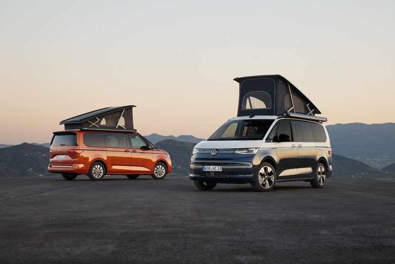 Dos furgonetas camper Volkswagen California con los techos elevables desplegados, estacionadas en un terreno elevado al atardecer o amanecer. Una furgoneta es de color naranja y la otra es bicolor (gris oscuro y blanco), con montañas en el fondo. La imagen evoca acampada y aventura en la naturaleza.